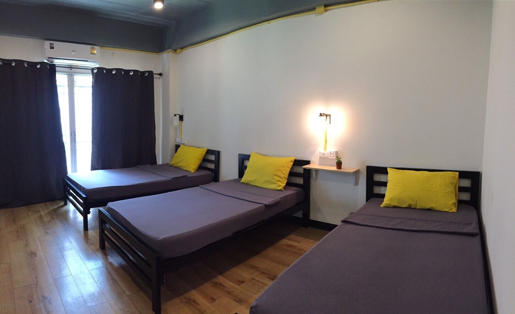 Une chambre avec trois lits simples, chacun doté d’une tête de lit et d’un oreiller. On y trouve une petite étagère avec une lampe et une plante.