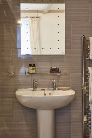 A bathroom with a white sink, two faucets, a mirror with lights, a glass shelf with toiletries, and a towel rack. - Lyme Townhouse (Lyme Regis)