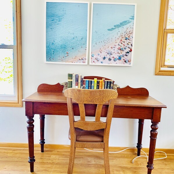 Un escritorio de madera con una silla, una estantería llena de libros y dos cuadros con motivos playeros en la pared.