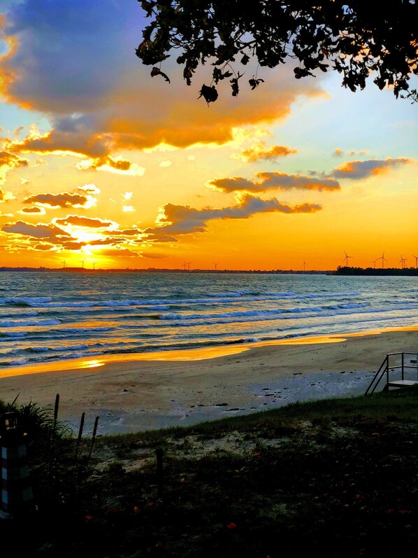 Una playa con atardecer, olas y aerogeneradores a lo lejos.
