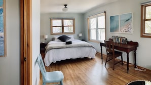 A bedroom with a bed, bedside tables, a desk, and a chair. There are windows, a ceiling fan, and artwork on the walls.