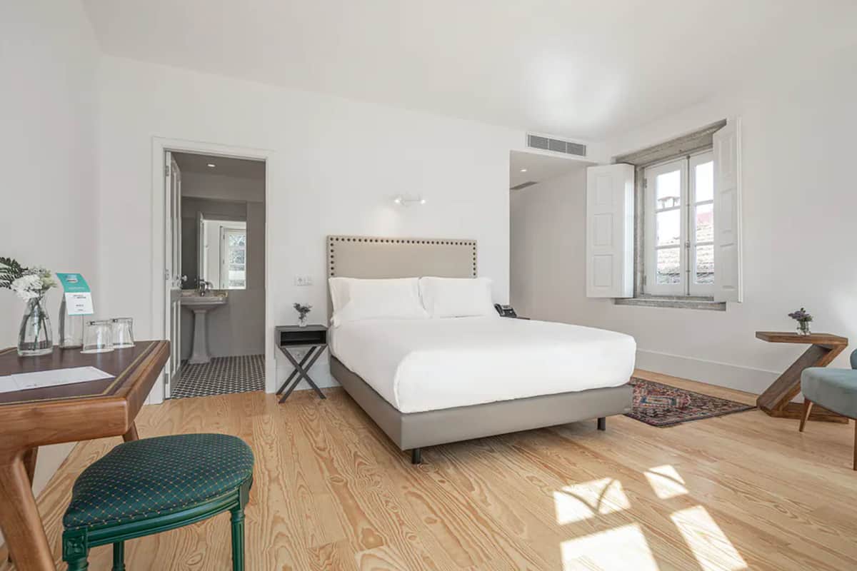 Quarto de hotel moderno com uma cama grande, uma escrivaninha com cadeira, um banheiro pequeno e uma janela com vista.