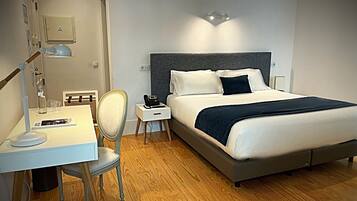 Una habitación de hotel ordenada con cama, mesita de noche, lámpara, sillón y escritorio con silla.