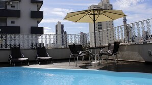 Un espace piscine aménagé sur le toit, avec des chaises longues et un parasol, entouré d’une rambarde décorative et de bâtiments en arrière-plan.