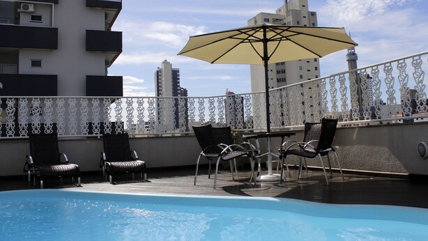 Un área de piscina en la azotea con sillones de descanso y una sombrilla, rodeada por una baranda decorativa y edificios al fondo.