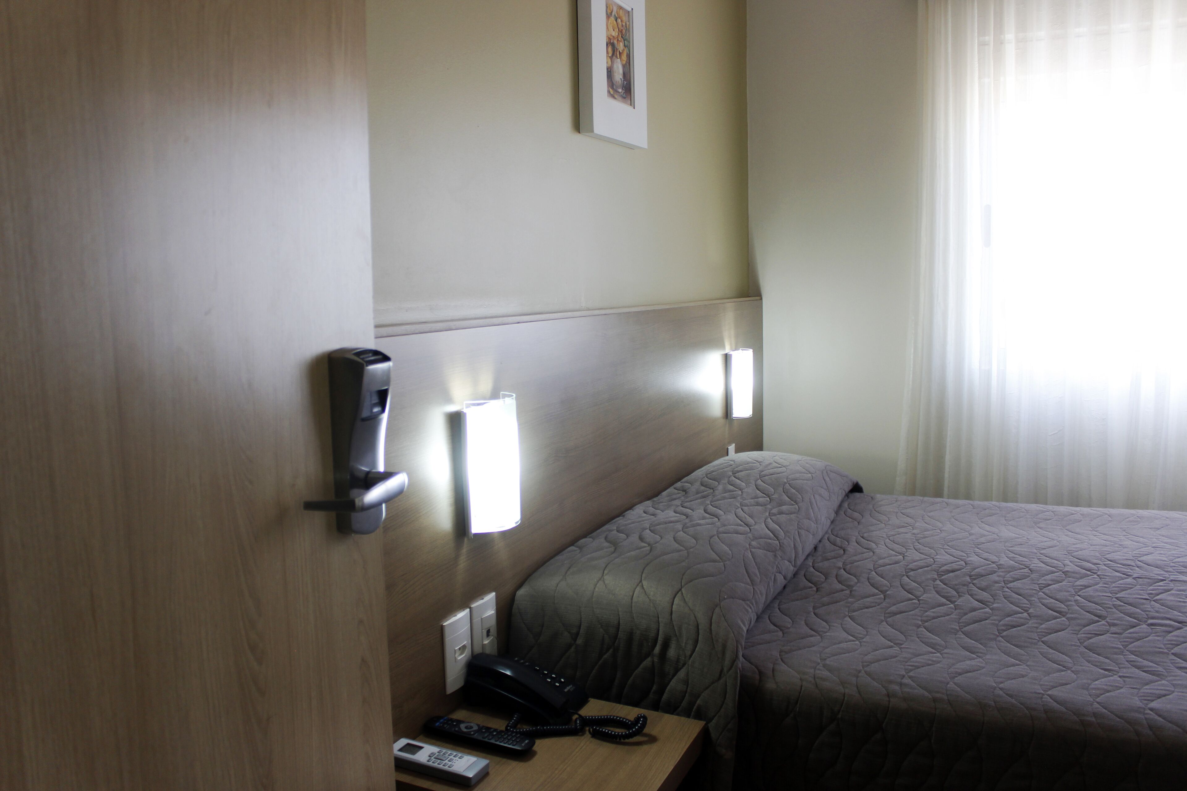 A hotel room with a bed, a bedside table with a phone and remote control, a wall-mounted light, and a picture on the wall.