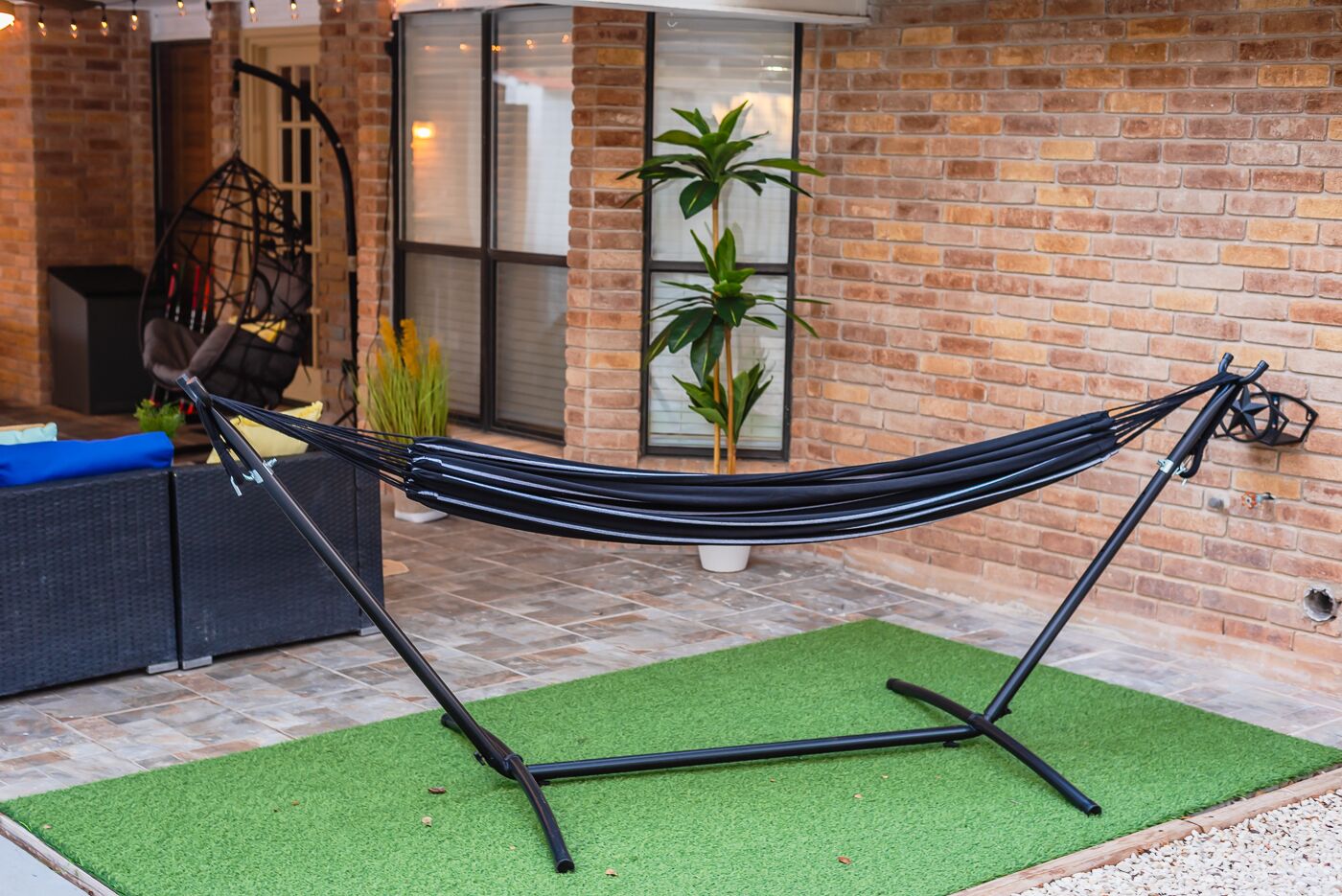 A hammock set up on a green artificial turf in front of a brick building with a glass door and potted plants.