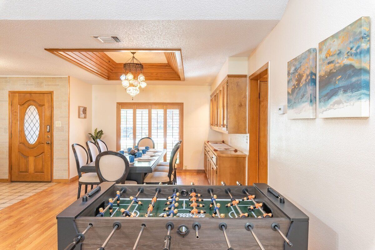 A room with a foosball table, wooden ceiling beams, a chandelier, a dining table with chairs, and a door with a glass panel.