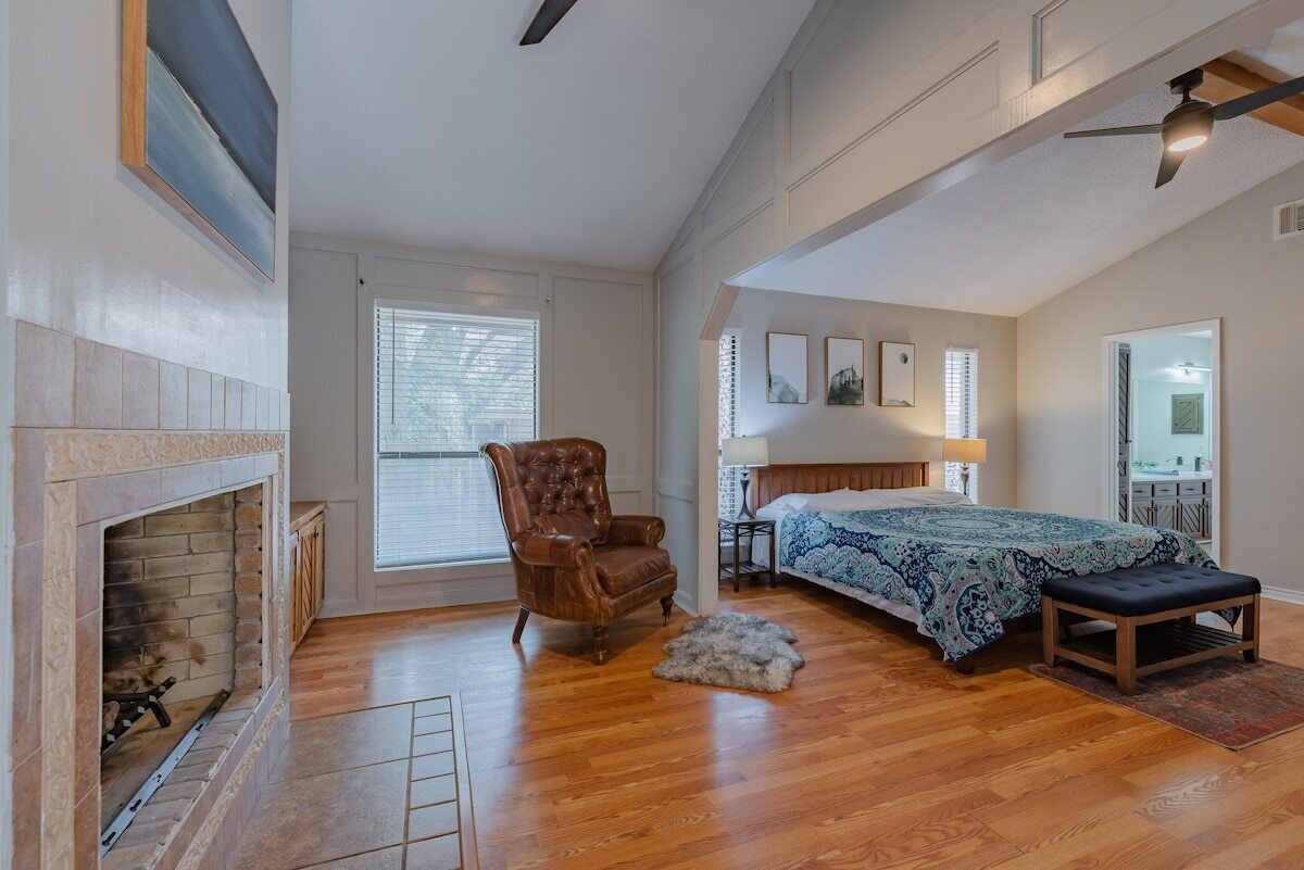 A bedroom with a fireplace, a bed with a patterned bedspread, a wooden floor, a leather armchair, and a ceiling fan.