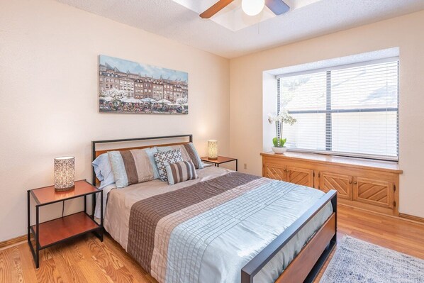 Une chambre à coucher avec un lit, une table de chevet, une lampe, une fenêtre avec des stores, un banc en bois et un tableau accroché au mur.