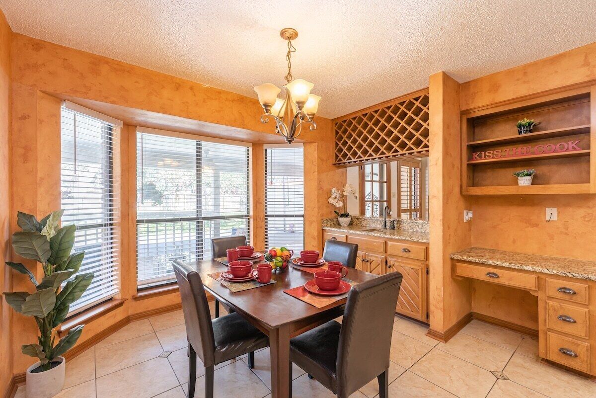 Comedor con mesa de madera preparada para cuatro personas, una araña y zona de cocina con armarios de madera y un letrero que dice 'KISS THE COOK'.