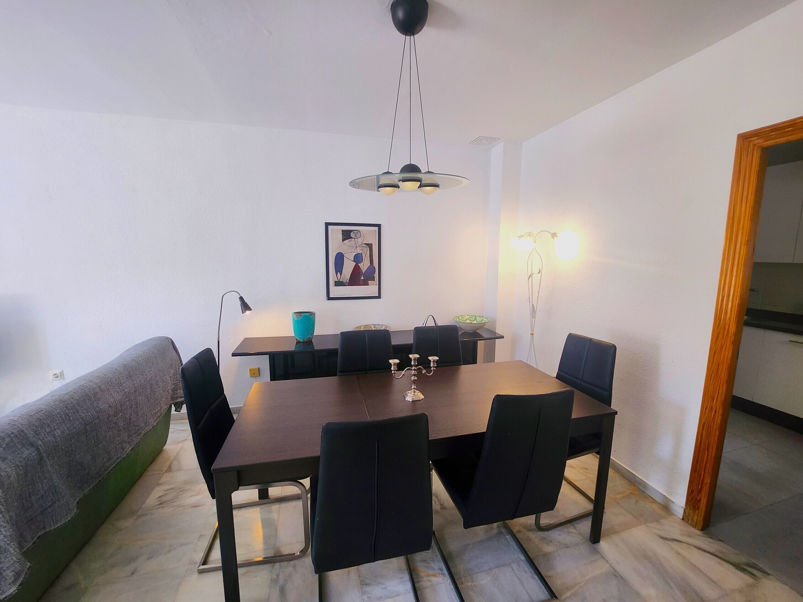 A dining area with a rectangular table, black chairs, a blue vase, a framed picture, and a pendant light.