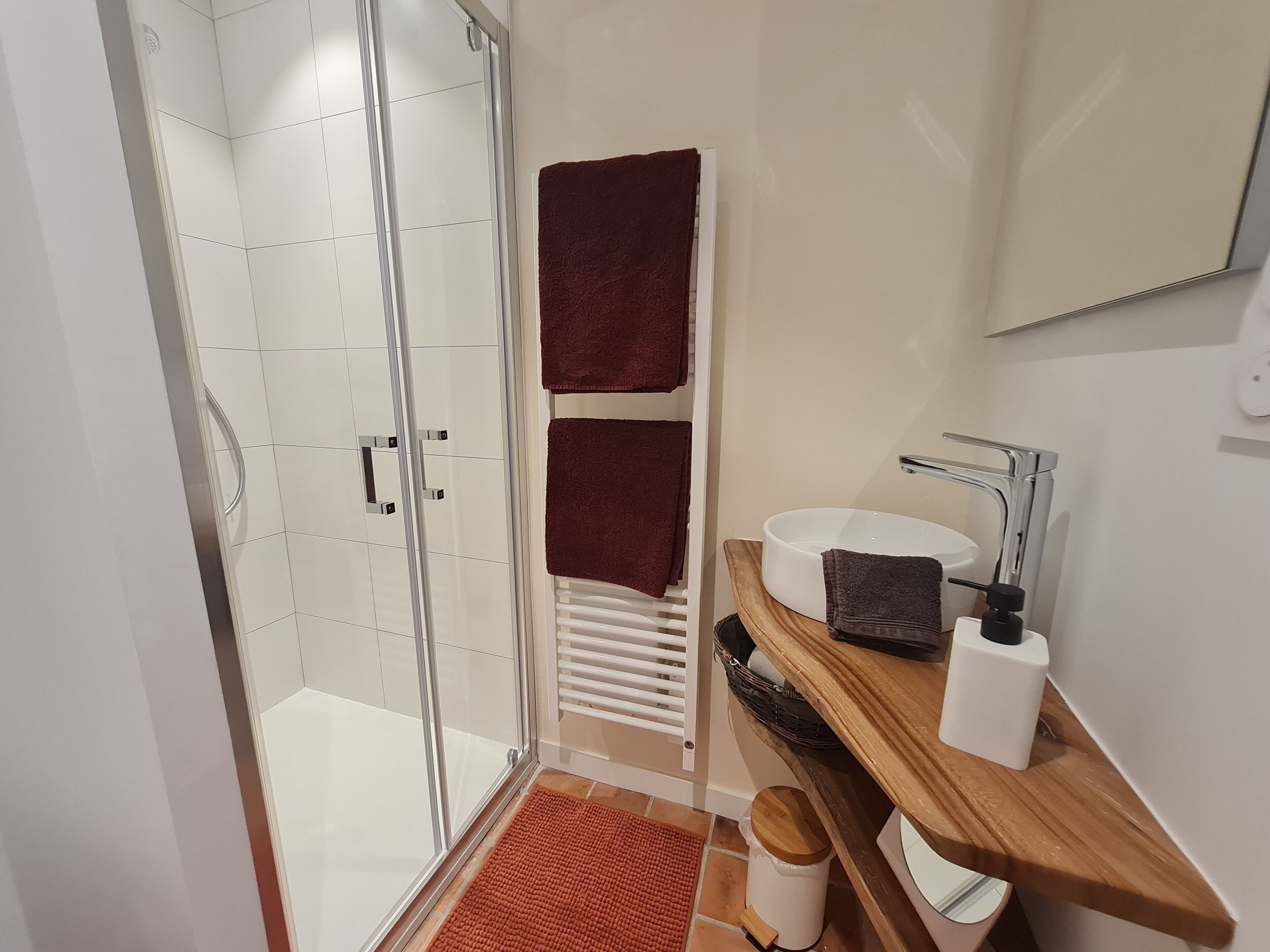A modern bathroom with a glass shower enclosure, a white sink with a wooden vanity, and two dark towels on a rack.
