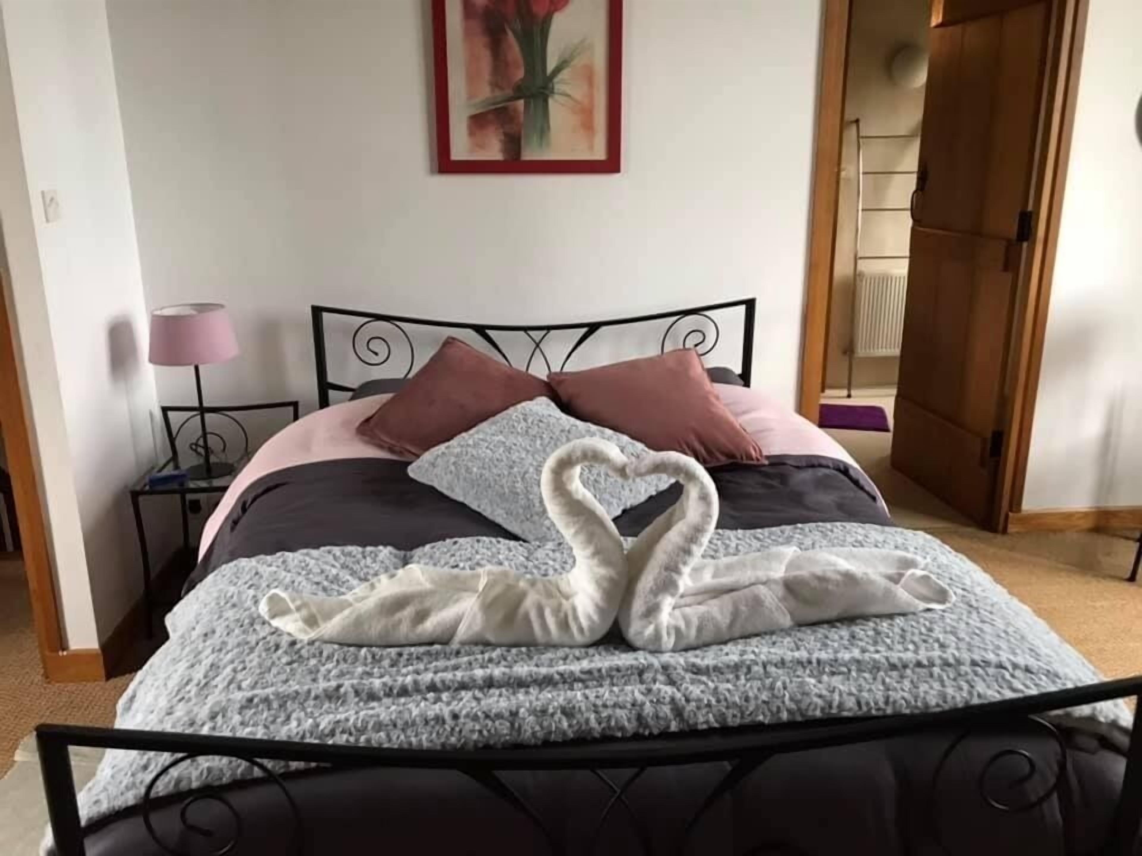 A neatly made bed with a heart-shaped towel arrangement, a lamp, and a painting on the wall.