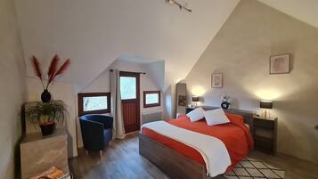 Une chambre avec un plafond en pente, un lit avec une literie blanche et orange, une chaise bleue, une table de chevet avec une lampe et une fenêtre donnant sur des arbres.
