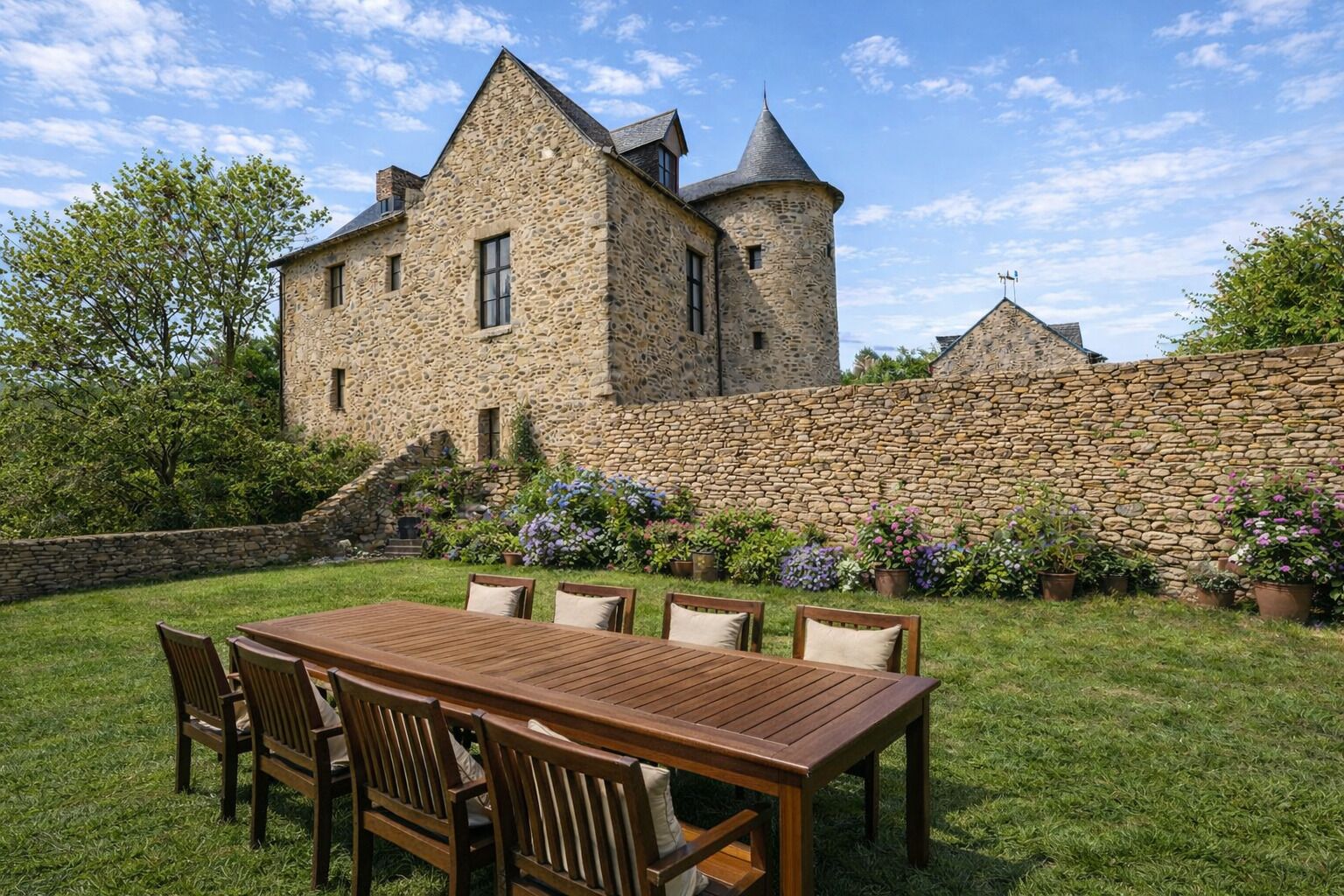 A stone building with a tower, surrounded by a stone wall and a grassy area with a wooden table and chairs.