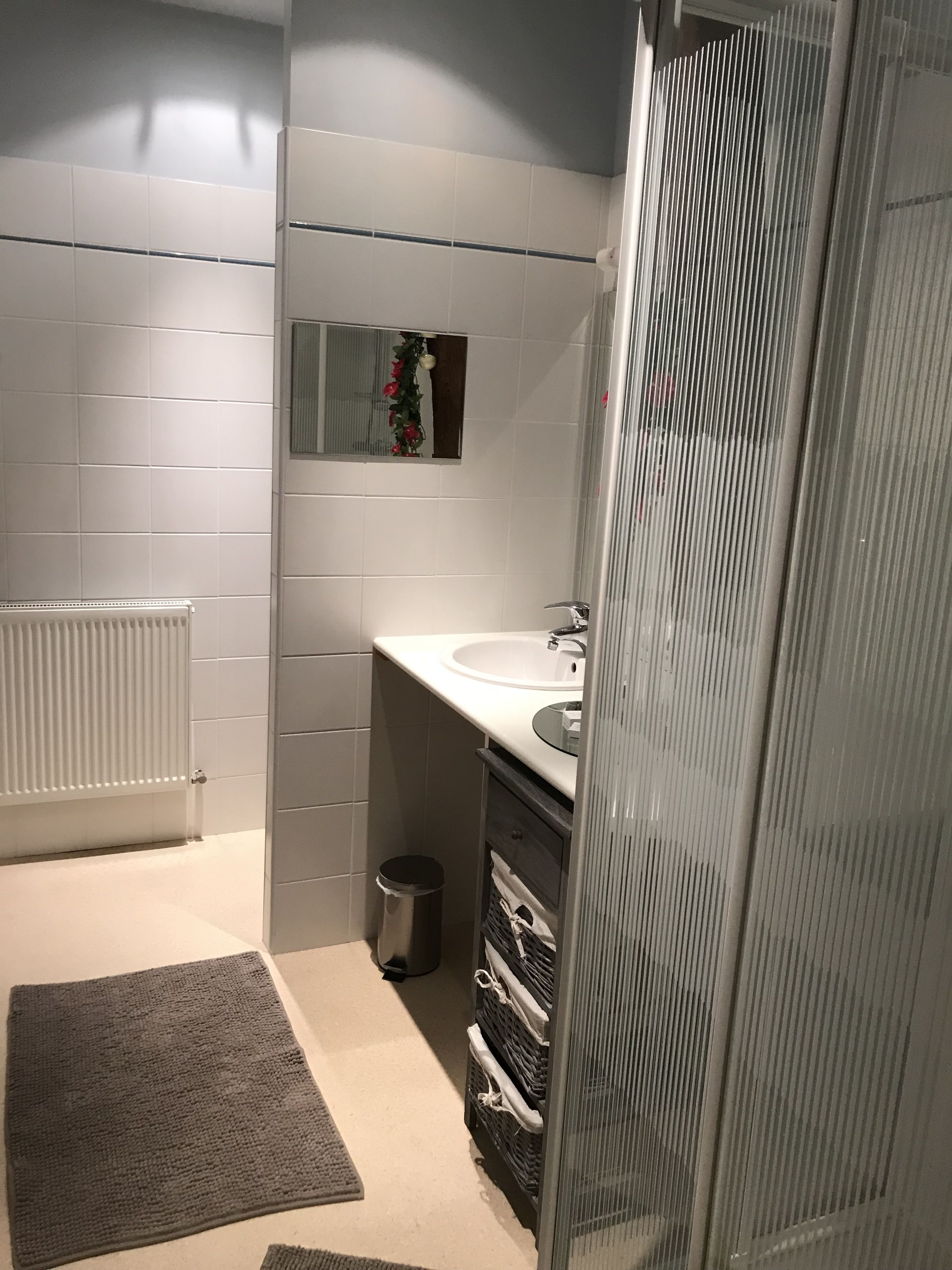 A bathroom with white tiles, a white sink, a mirror, a radiator, a trash bin, and a shower area.