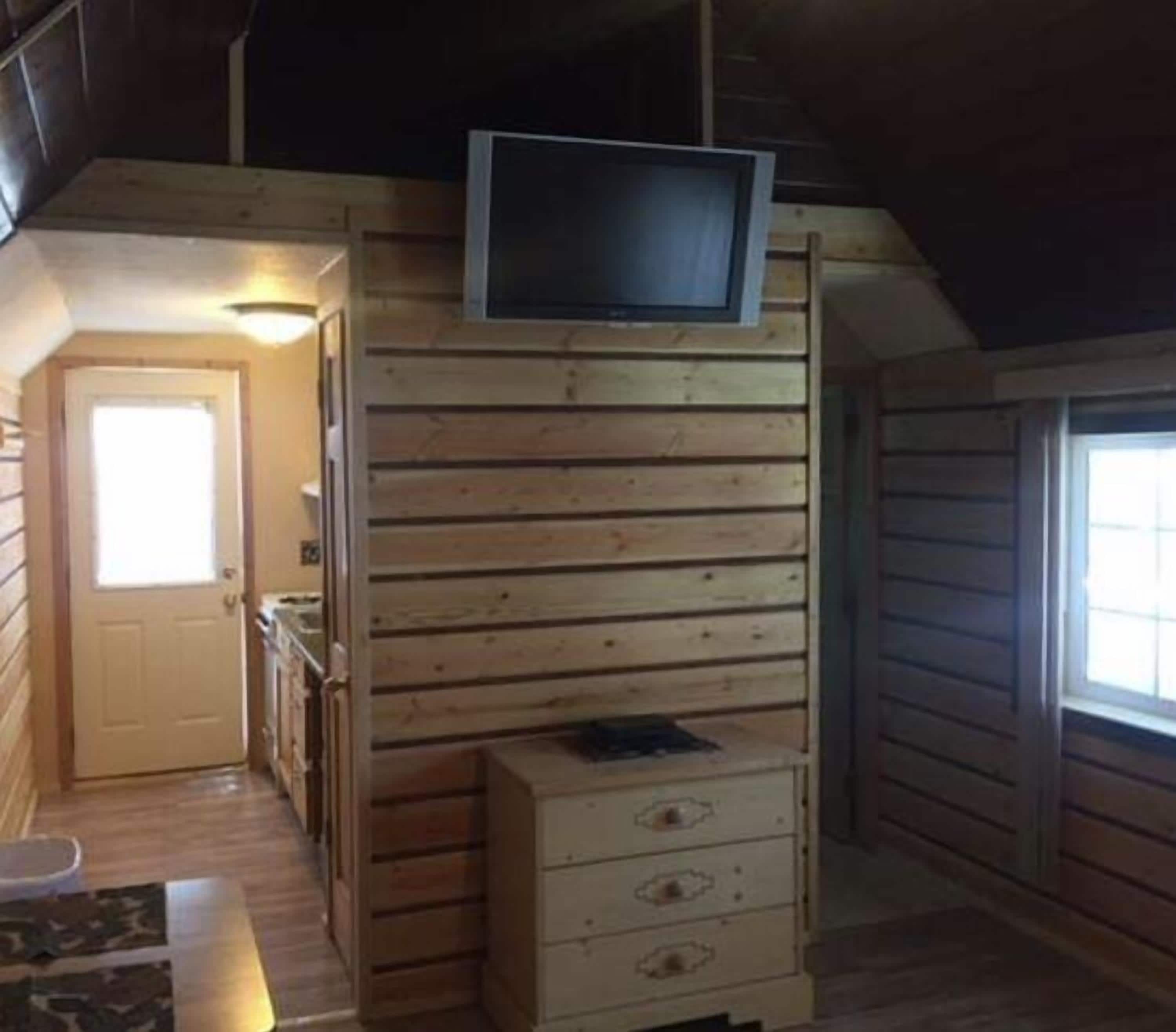 A wooden cabin interior with a television mounted on the wall, a small wooden dresser, and a door leading to another room.