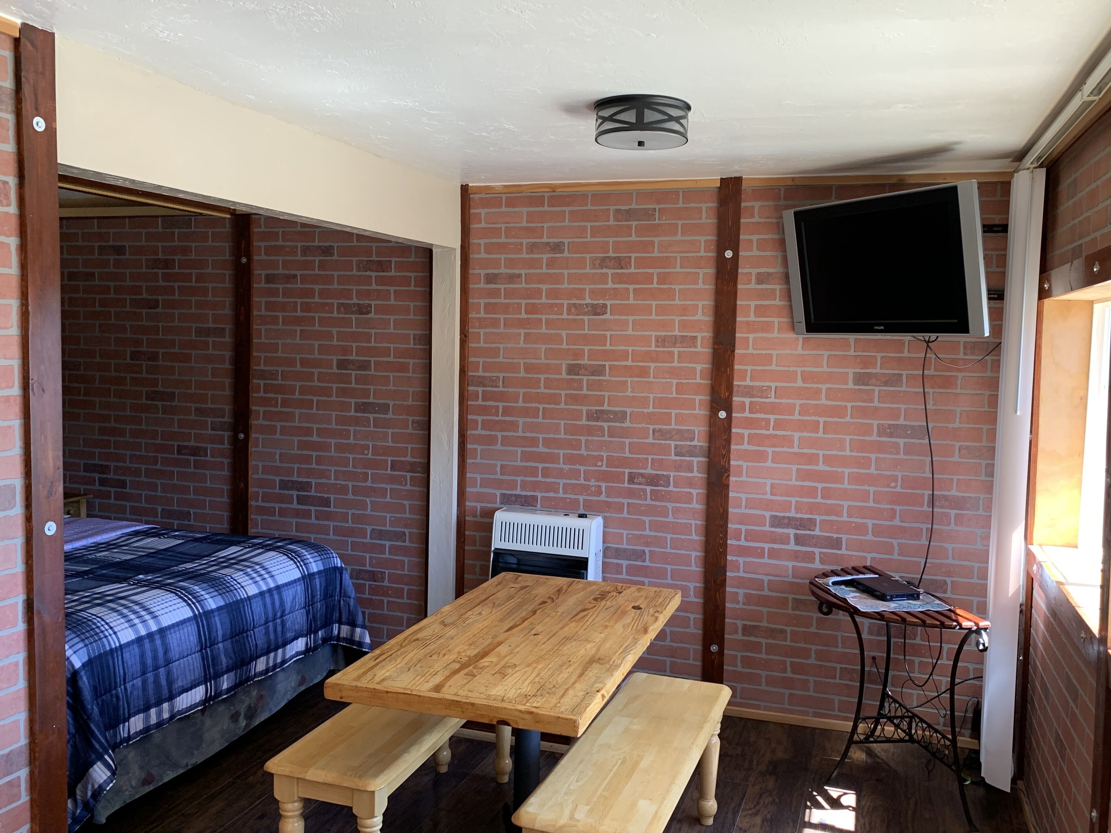 Habitación compacta con cama, una mesa de madera con bancos, televisor montado en la pared y radiador.