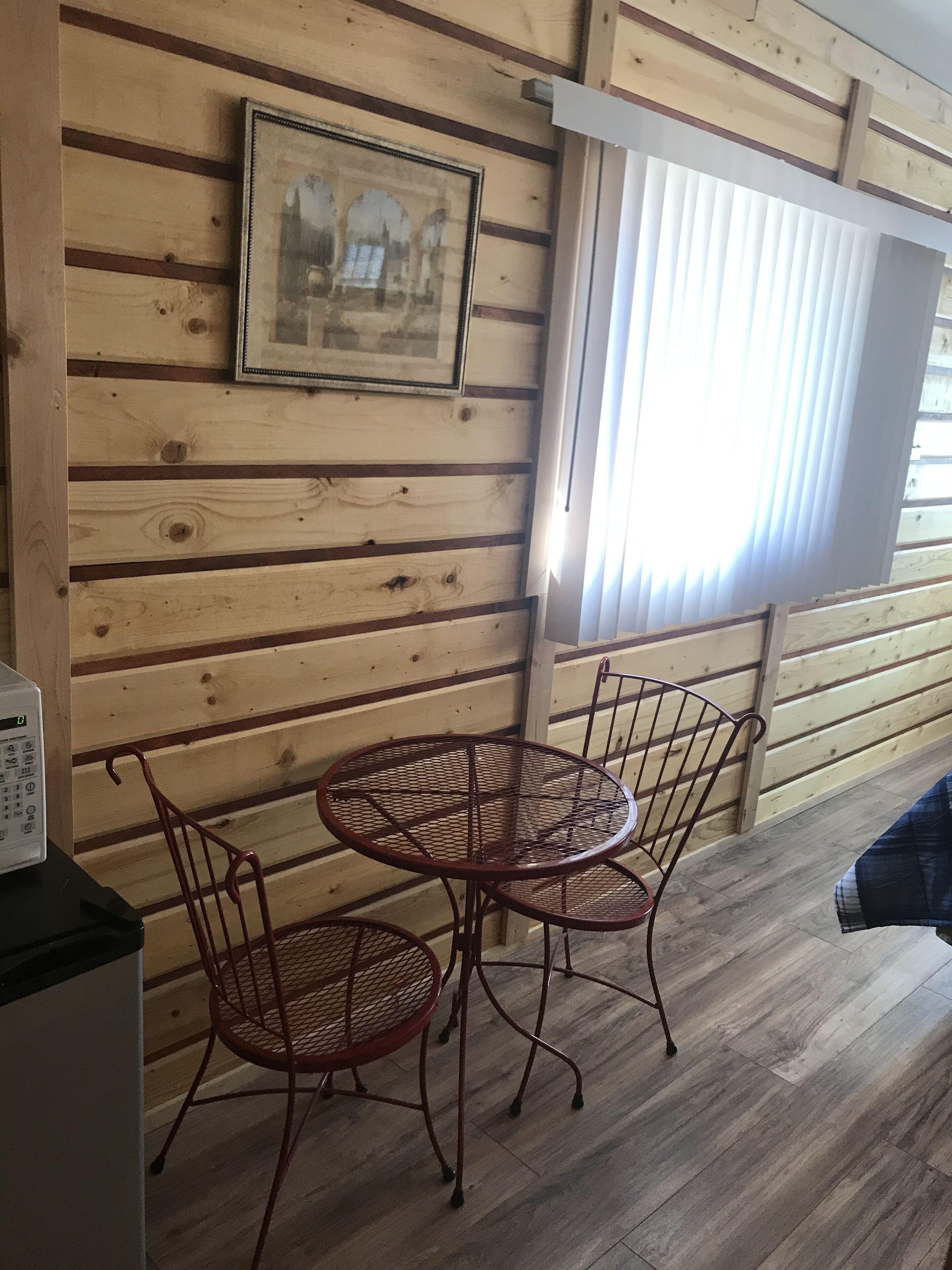 A wooden interior with a round metal table and chairs, a framed picture on the wall, and a window with blinds.