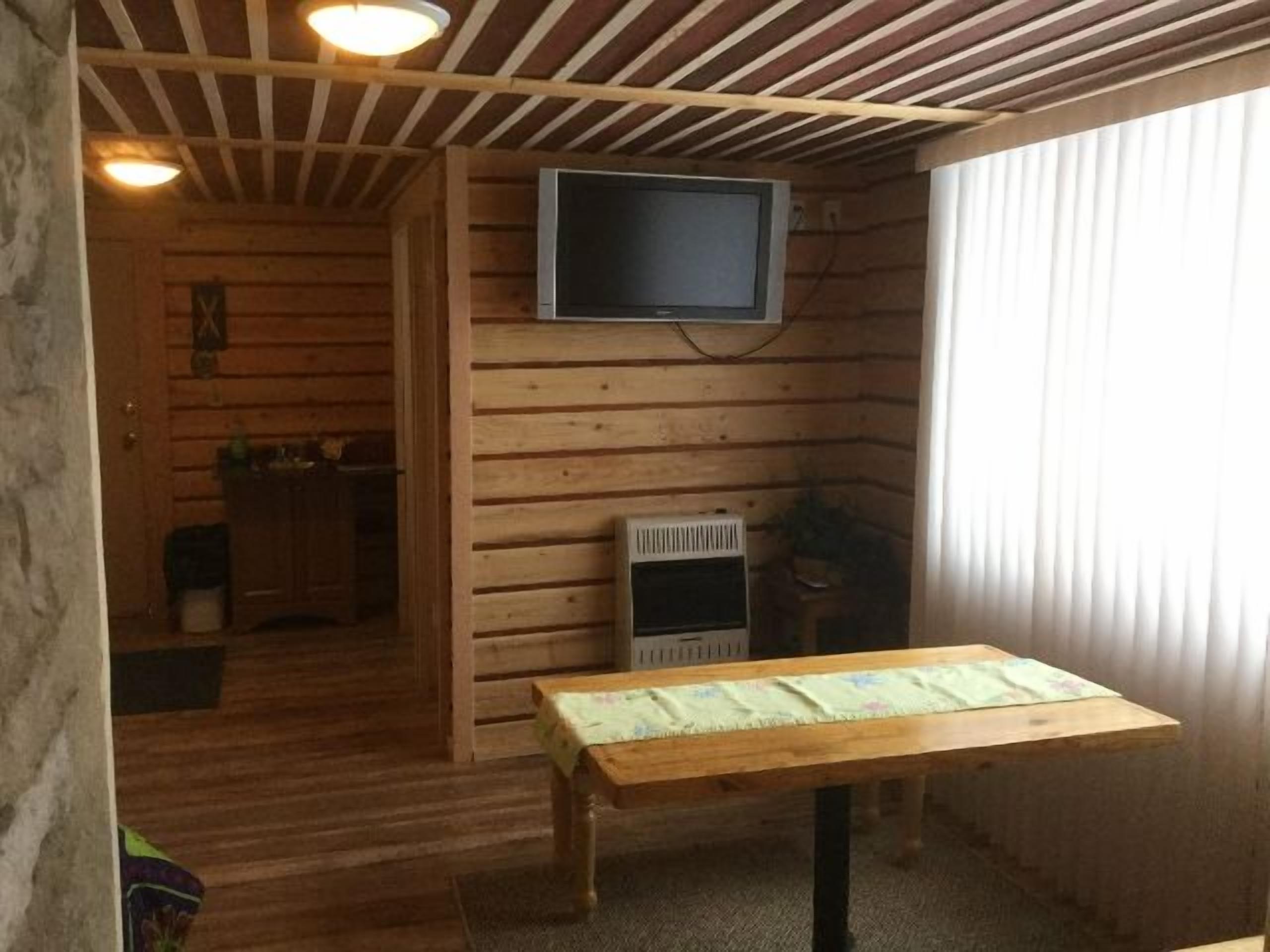 A cozy room with wooden walls and a ceiling, a flat-screen TV mounted on the wall, a wooden table with a patterned tablecloth, and a window with curtains.