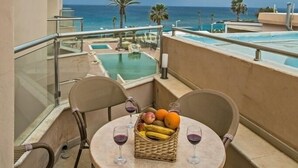 Un balcon avec une table dressée pour deux personnes, un panier en osier rempli de fruits et des verres à vin, et une vue sur l’océan.