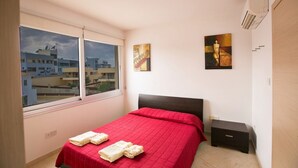 Une chambre avec une grande fenêtre, un lit avec une literie rouge, deux tableaux accrochés au mur et une petite table de chevet.