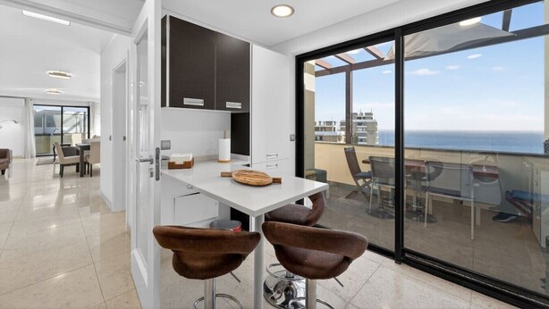 A modern kitchen with a dining area, large windows, and a view of the sea.