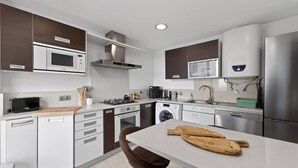 A modern kitchen with white cabinets, stainless steel appliances, a built-in oven, microwave, and dishwasher. There is a wooden cutting board on the counter and a brown chair.