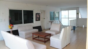 A spacious living room with a large window, white sofas, a wooden coffee table, and a small side table with a vase of flowers. - OYO Hotel Colibri (Chetumal)