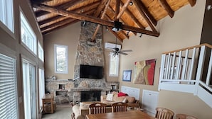 A spacious living room with a stone fireplace, wooden ceiling beams, and a large dining table.