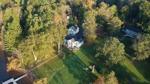 An aerial view of a large house surrounded by dense forest, with a visible driveway and a dock by a body of water.