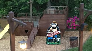 A wooden play structure with a slide and a platform, surrounded by greenery and a pink flower bed.