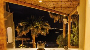 Zona junto a la piscina con sillas de mimbre, una mesa con un vaso y vistas a palmeras de noche.