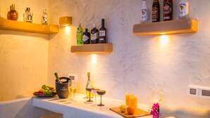 Zona de bar con estantes llenos de botellas de alcohol, un cubo con abrelatas, vasos y un frutero.