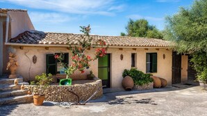 A traditional Mediterranean-style building with a tiled roof, a stone wall, and a garden area with potted plants and a statue. - Small, cozy.  App. Petit der FincaOase- rental -ONLY ADULD (Manacor)