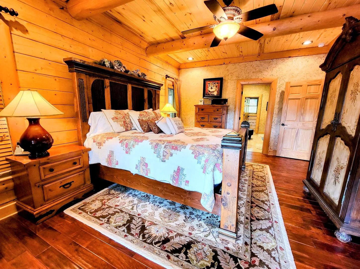 A wooden bedroom with a bed, nightstands, a lamp, a ceiling fan, and a door.