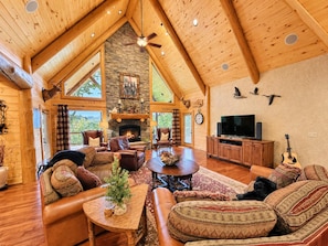 A spacious living room with a stone fireplace, leather furniture, a television, and a guitar. - Luxury Mountain Lodge | Big Views, Game Room, Hot Tub, Fire Pit & Paved Access (Cherry Log)