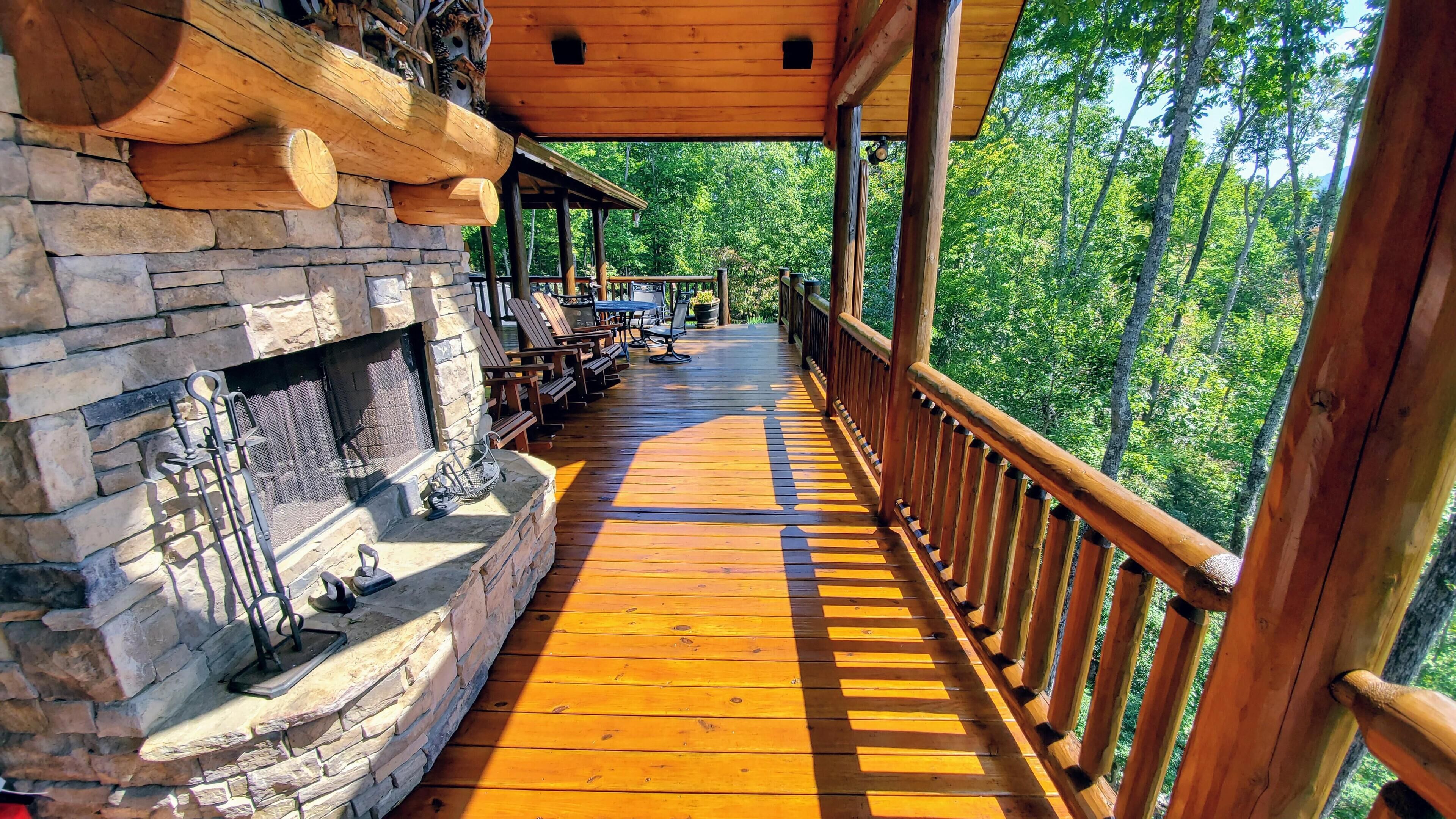 A wooden deck with a stone fireplace, outdoor seating, and a view of the forest.