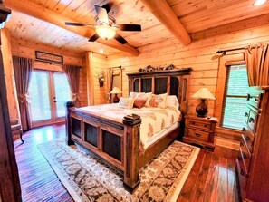 A wooden cabin bedroom with a large bed, a ceiling fan, a dresser, and a window with curtains. - Luxury Mountain Lodge | Big Views, Game Room, Hot Tub, Fire Pit & Paved Access (Cherry Log)