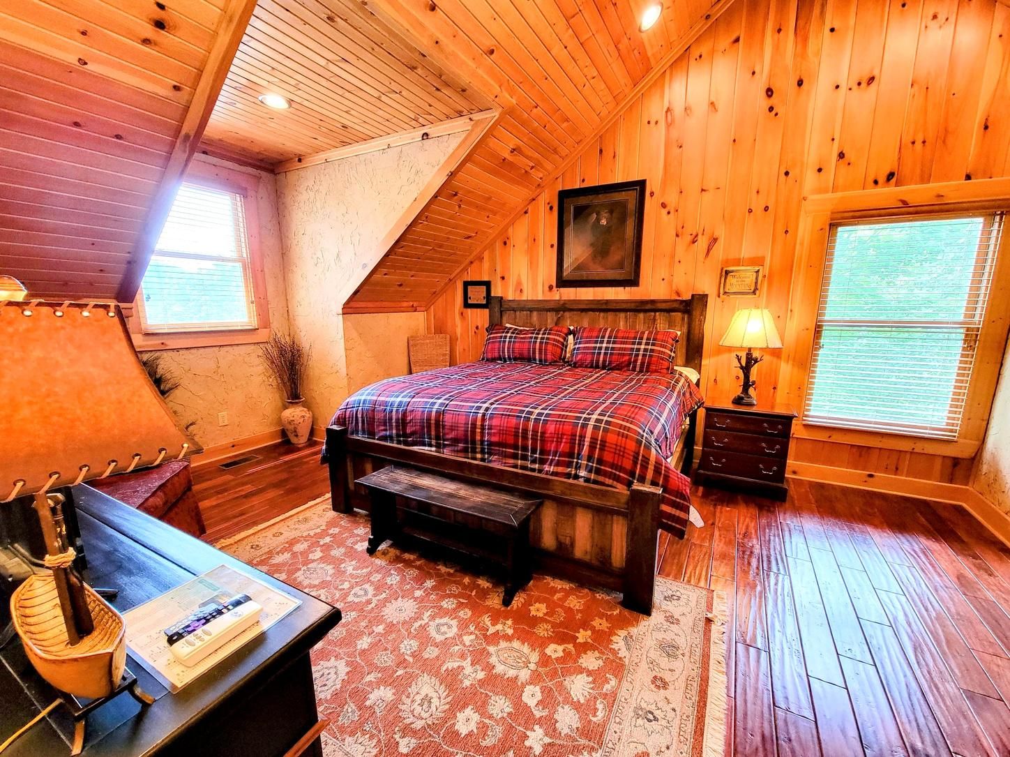 A bedroom with a wooden ceiling, a bed with plaid bedding, a nightstand, a lamp, and a window with blinds.