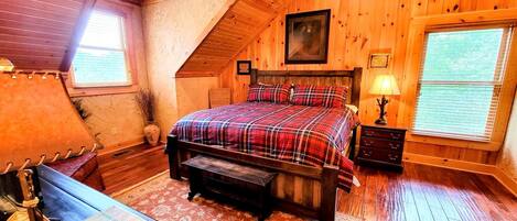 A bedroom with a wooden ceiling, a bed with plaid bedding, a nightstand, a lamp, and a window with blinds.