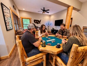 A group of people playing cards at a wooden table with poker chips, in a room with a ceiling fan, a mounted TV, and a wall with framed items.