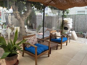 A covered outdoor seating area with wooden furniture and cushions, a potted plant, and a view of a tree and fence.