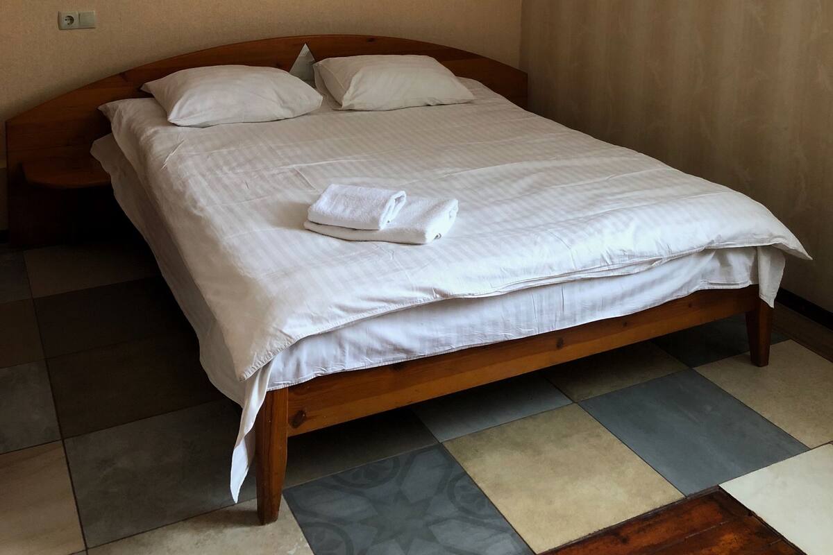 A neatly made bed with white linens and a folded towel, a wooden headboard, and a tiled floor with a mix of patterns.
