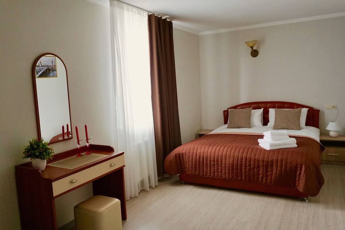 A hotel room with a bed, a desk with a mirror, a nightstand, and a wall-mounted lamp.