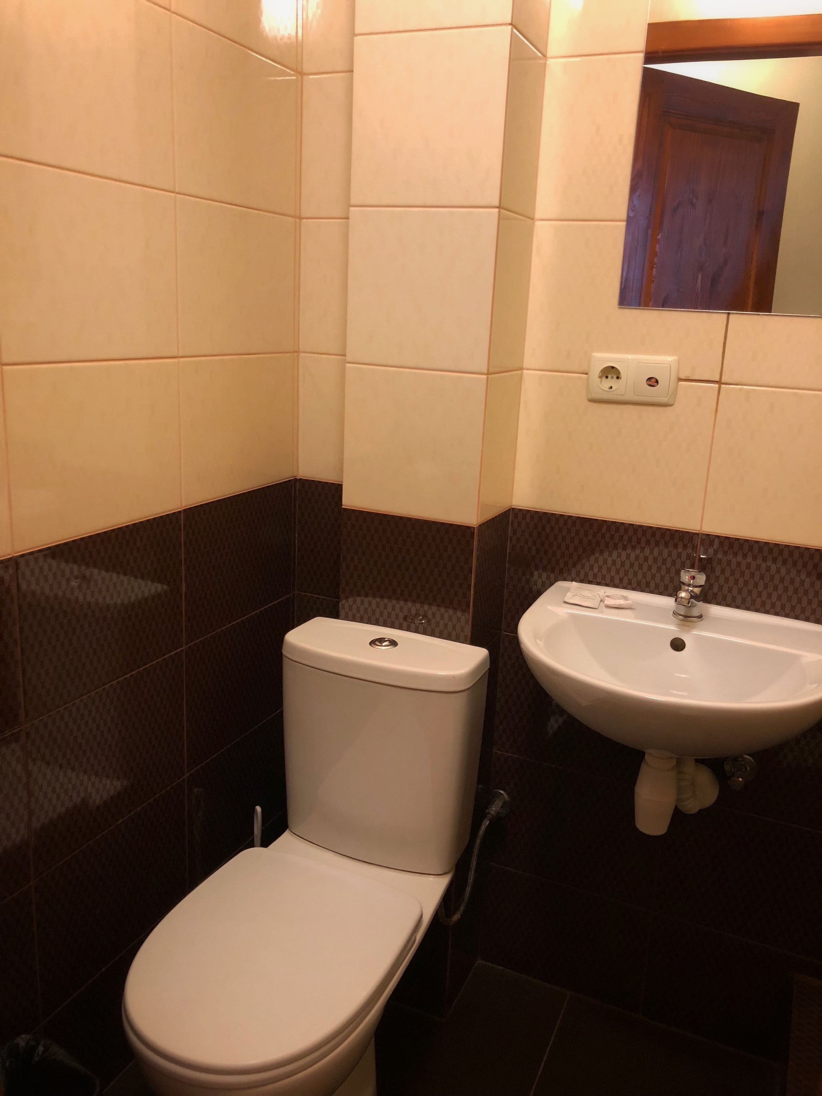 A bathroom with a toilet and a sink, both in white, against a backdrop of beige and dark brown tiles.