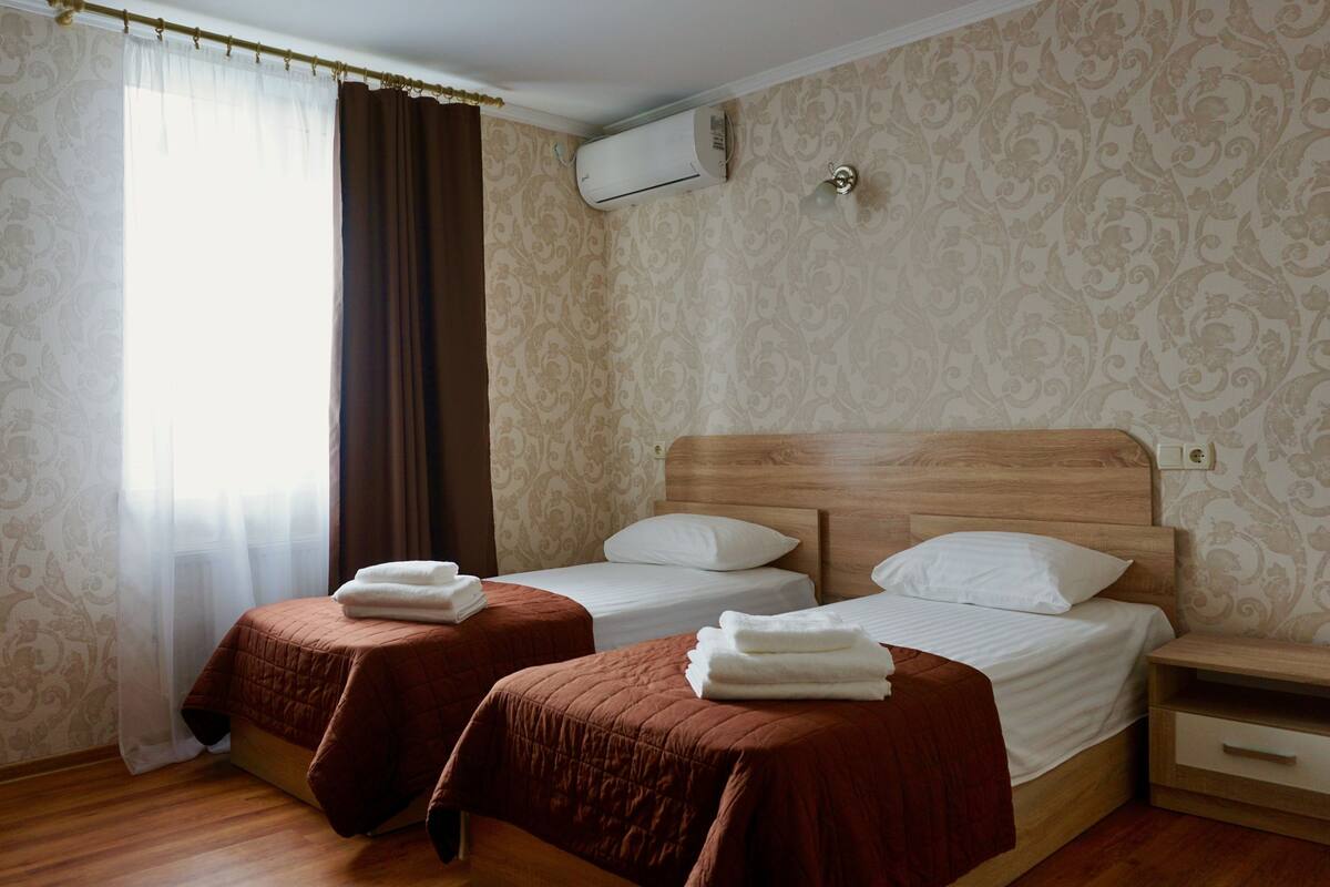 A hotel room with two beds, brown bedspreads, white pillows, a wooden headboard, a nightstand, and an air conditioning unit on the wall.