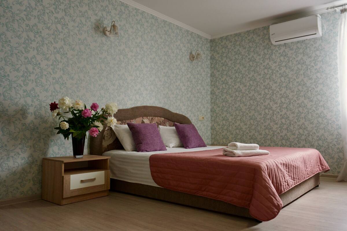 A neatly arranged bedroom with a wooden bed, a bedside table with a vase of flowers, and an air conditioning unit on the wall.