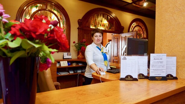 A hotel reception desk with a wooden counter, a computer, and a vase of flowers. - Hotel & Restaurant Zhuliany City (Sofiivska Borschahivka)